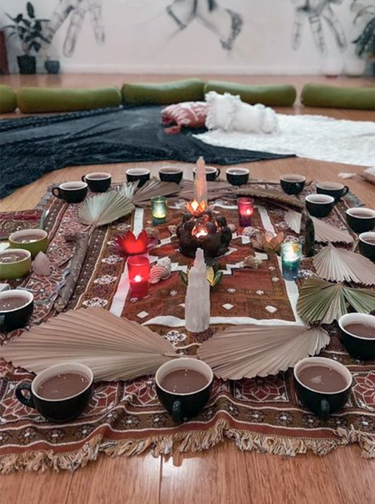 A traditional tea setup with cups and a central burner on an ornate rug in Sherman Oaks, surrounded by an arrangement of palm leaves and crystals.