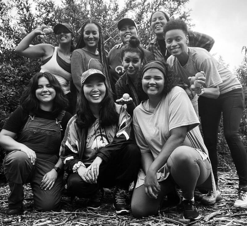 A group of nine people poses outdoors in front of their home. Some are standing, some squatting, and one making a muscle pose. They appear happy and are dressed in casual attire. Trees and a cloudy sky serve as the backdrop.